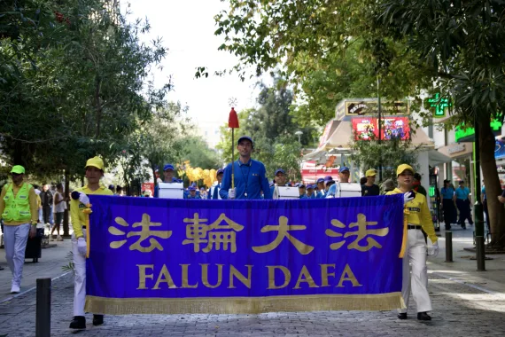 Falun-Dafa-Transparent an der Spitze der European Tian Guo Marching Band in einer baumgesäumten Straße der Athener Innenstadt, 2026