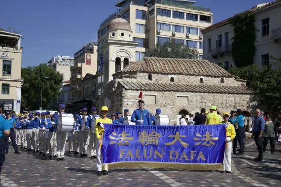 European Tian Guo Marching Band mit Falun-Dafa-Transparent vor der byzantinischen Pantanassa-Kirche am Monastiraki-Platz in Athen, 2026
