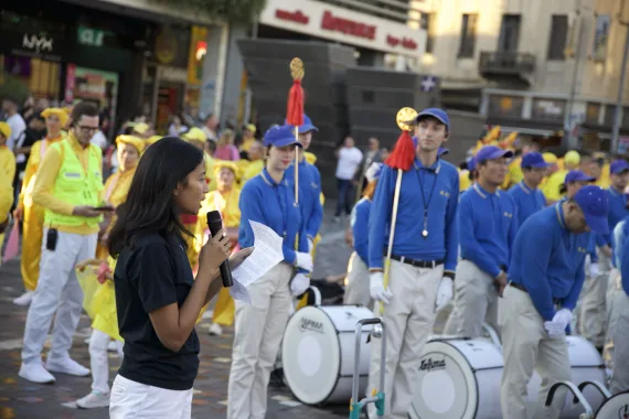 Eine Rednerin spricht mit Mikrofon zum Publikum, hinter ihr die European Tian Guo Marching Band mit ihren Trommeln, Athen 2026