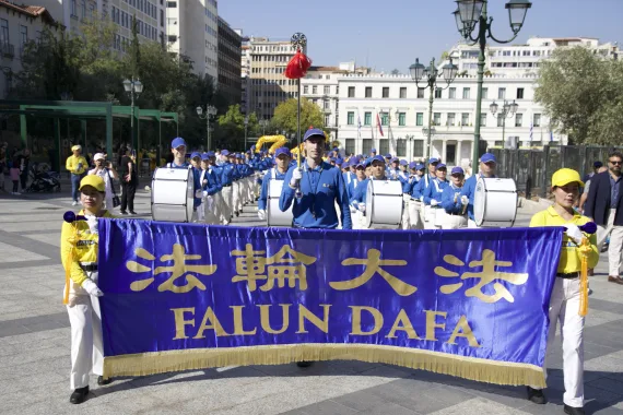 Tambourmajor und Trommler der European Tian Guo Marching Band hinter dem Falun-Dafa-Transparent am Syntagma-Platz in Athen, 2026