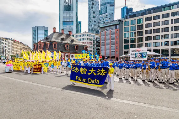European Tian Guo Marching Band mit Falun-Dafa-Transparent auf einem Platz vor der Skyline des Frankfurter Bankenviertels, 2026