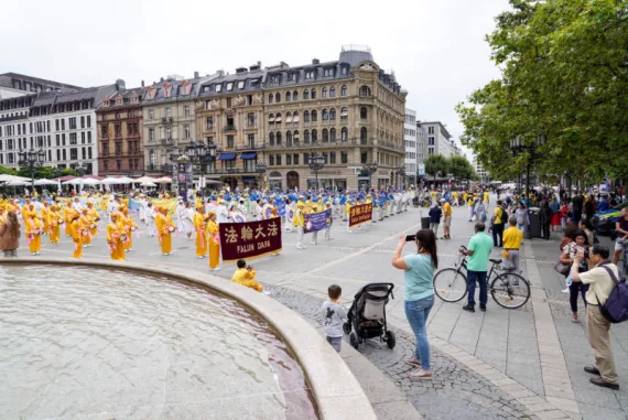 Falun-Dafa-Praktizierende in gelben Gewändern und die European Tian Guo Marching Band auf einem Platz mit Brunnen in Frankfurt am Main, 2021