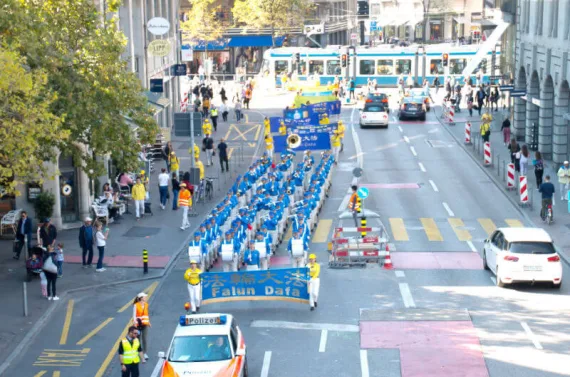 European Tian Guo Marching Band zieht mit Polizeibegleitung durch eine Innenstadt, im Hintergrund eine Straßenbahn