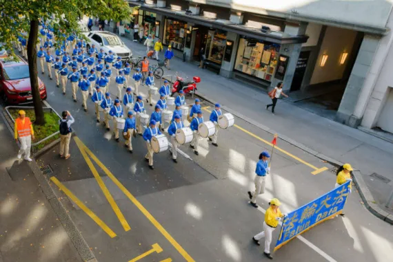 Blick von oben auf die European Tian Guo Marching Band, die mit Trommeln und Falun-Dafa-Banner durch eine Geschäftsstraße zieht